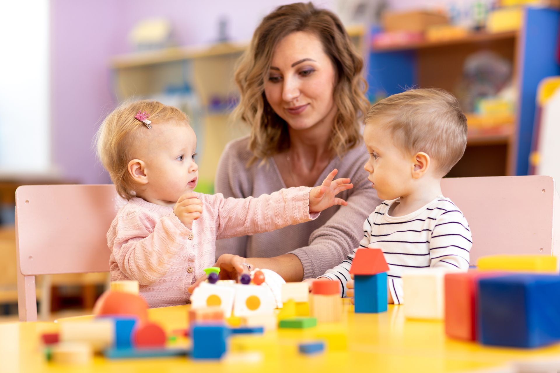 View of 2 babies with a teacher in preschool playing with toys.