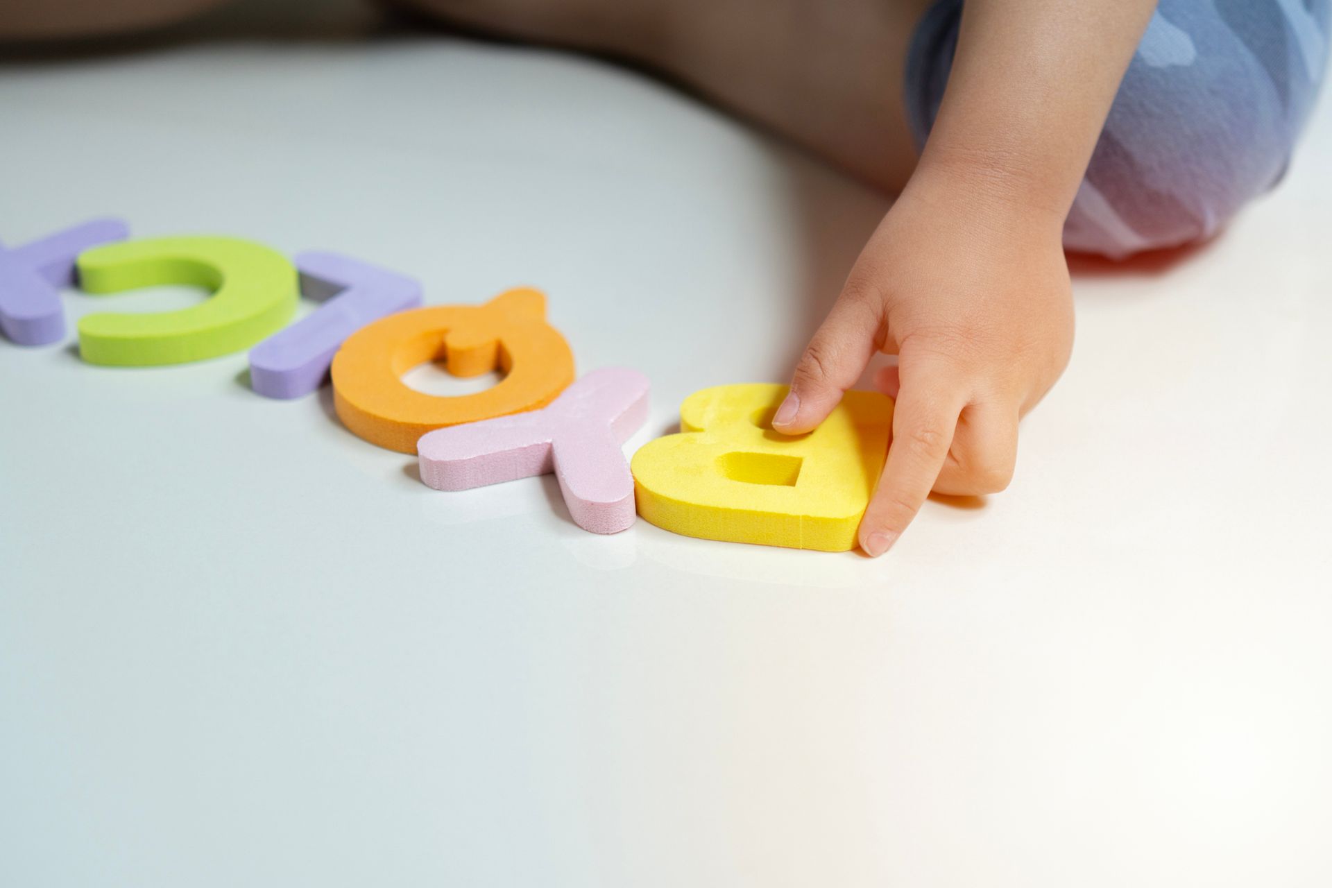 Child's hand playing with colourful foam letters on white surface.