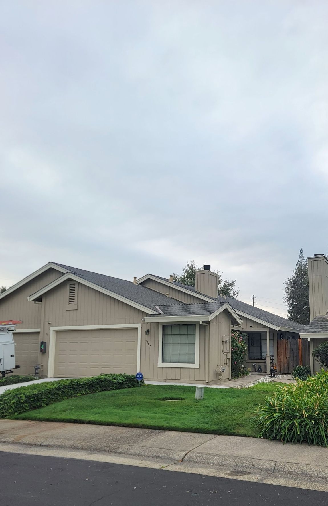 Tan house with gray roof, chimney, garage, and front yard with green grass and hedges under a cloudy sky.