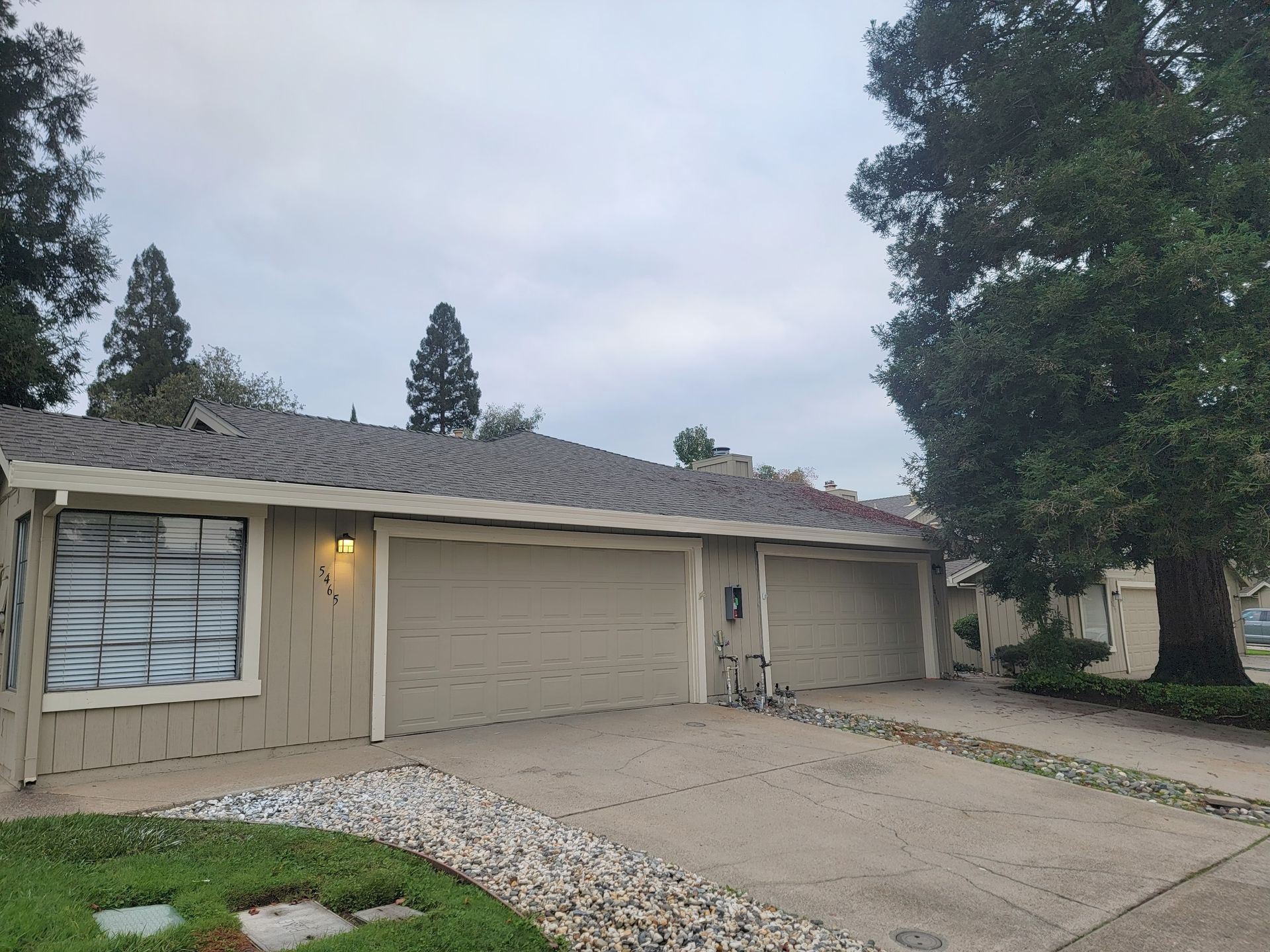 Beige home with two garage doors, concrete driveway, and a large tree under a cloudy sky.