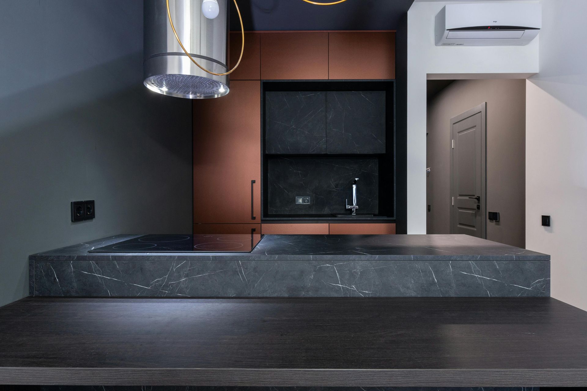 Modern kitchen featuring a black marble island, copper cabinets, a stainless steel range hood, and a doorway.