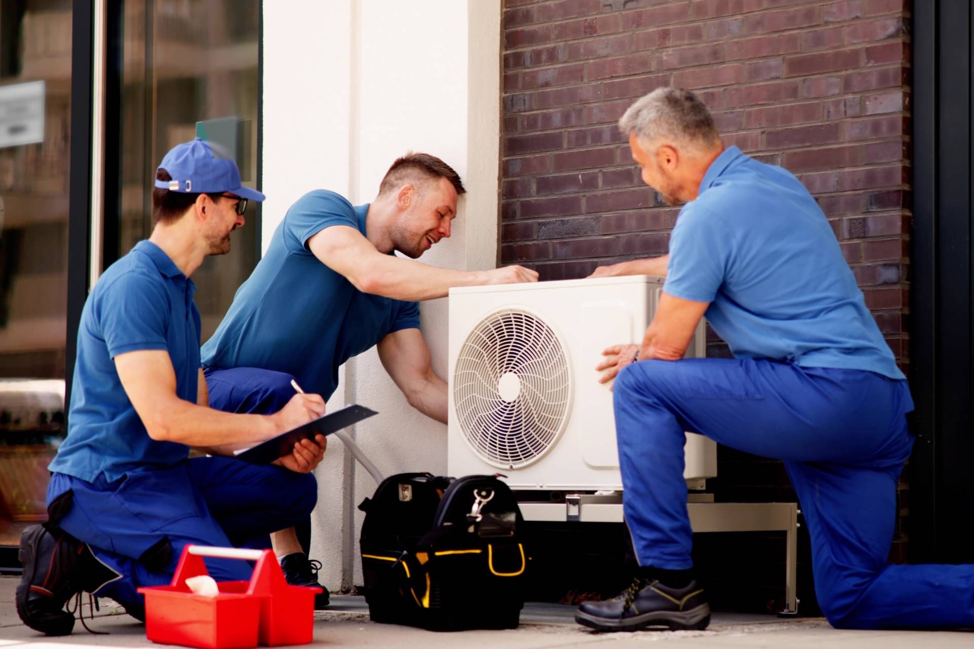 Three HVAC technicians in blue uniforms install an air conditioning unit outside.