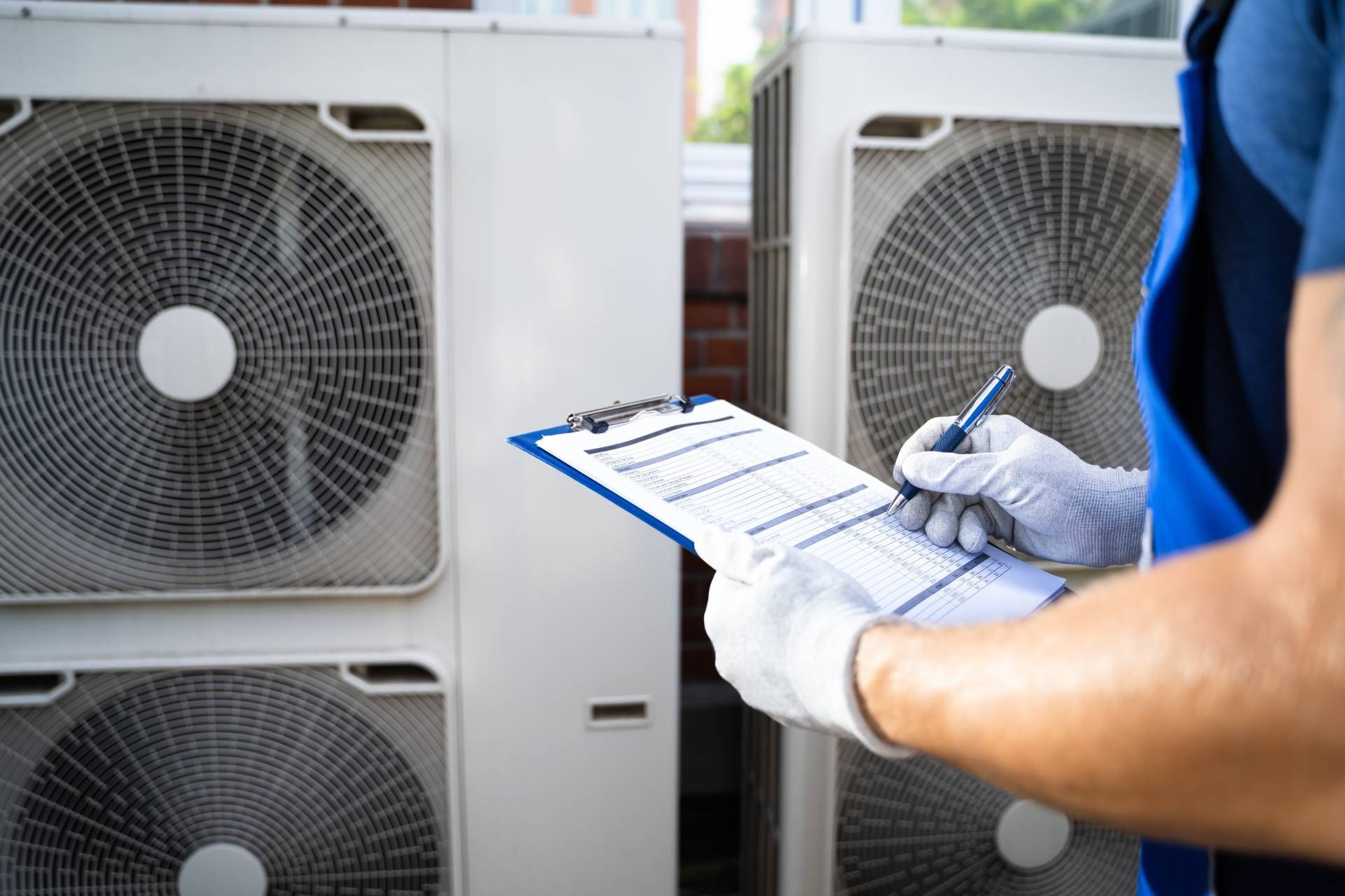 HVAC technician in blue uniform and gloves, inspecting outdoor air conditioning units with clipboard.