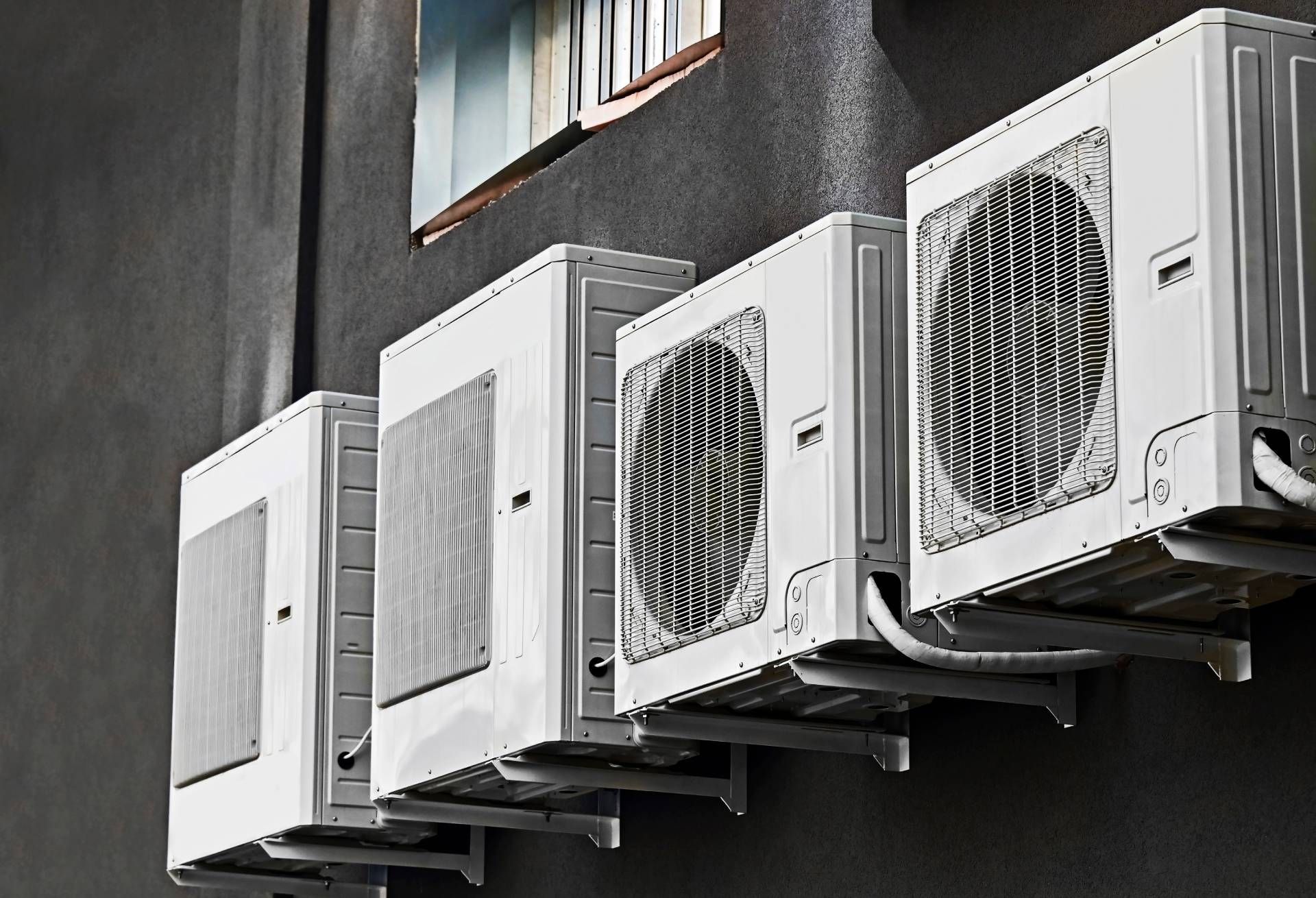 Four white air conditioning units mounted on a dark gray building wall, near a window.