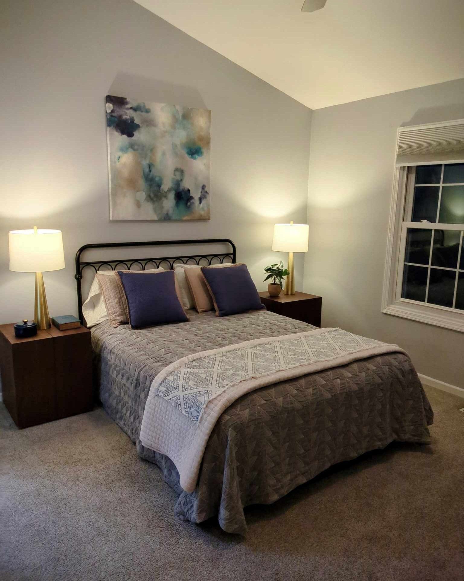 Bedroom with gray walls, bed with blue pillows and a quilt, nightstands, lamps, and a painting.