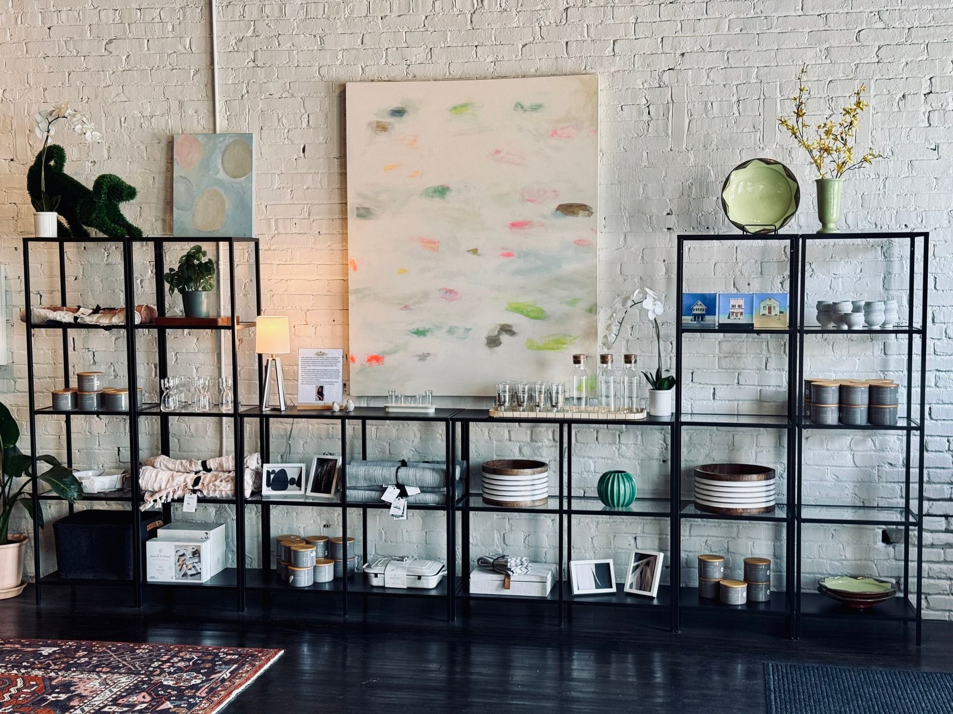 Shelving unit display in a shop with home decor: art, vases, candles, and plants against a brick wall.