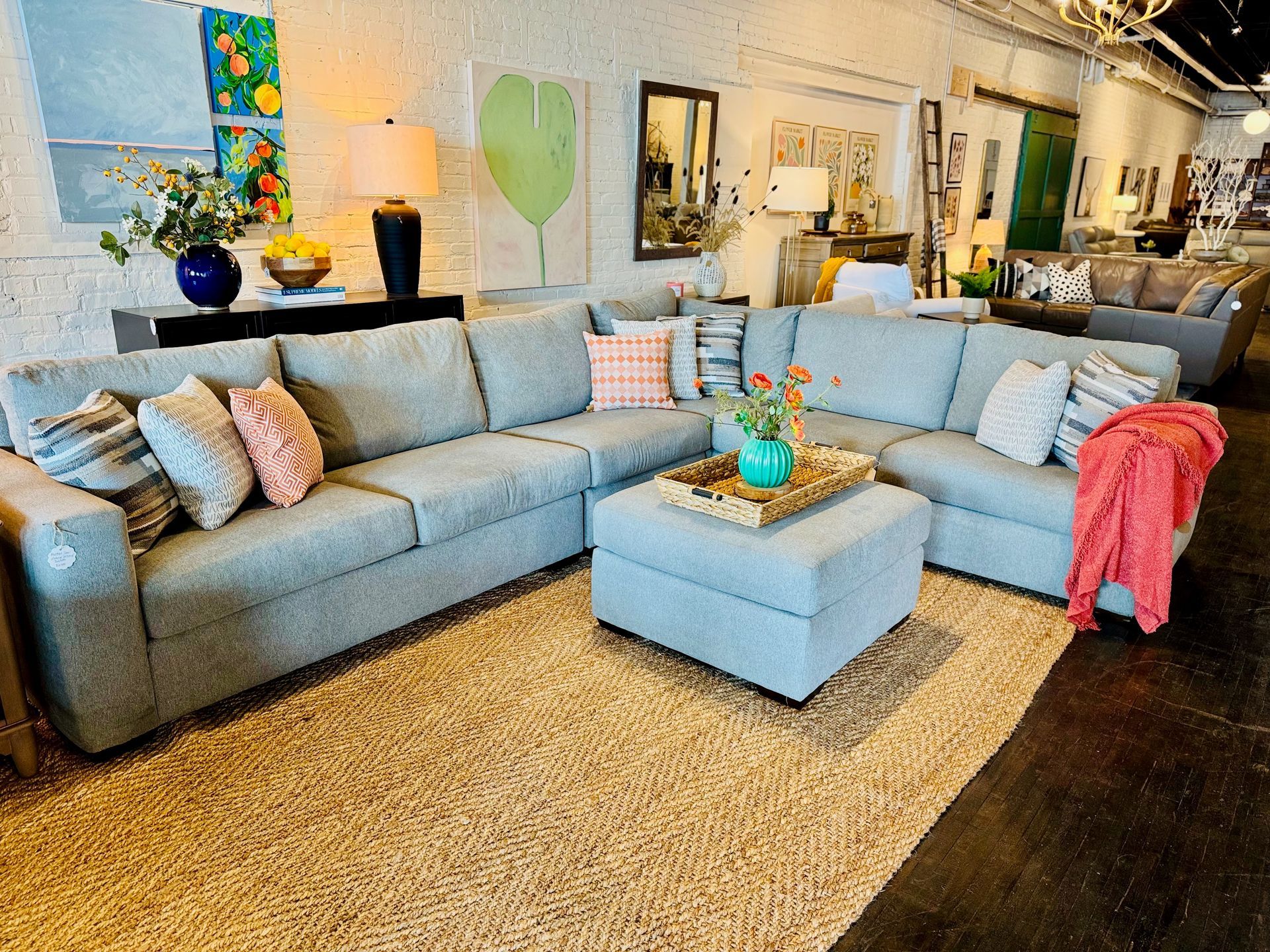Blue sectional sofa and ottoman with pillows and blanket in a brightly lit room, on a woven rug.