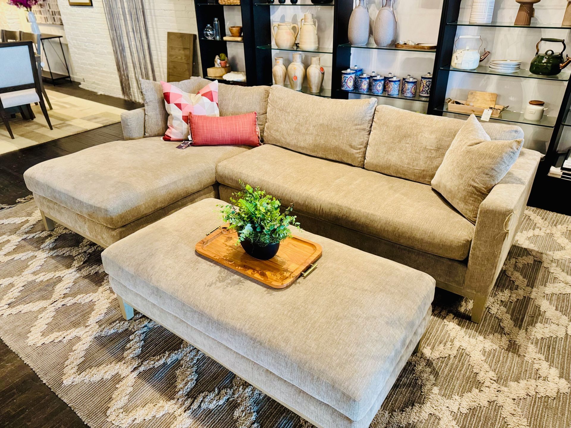Beige sectional sofa with ottoman and throw pillows in a showroom, with decor on shelves in the background.