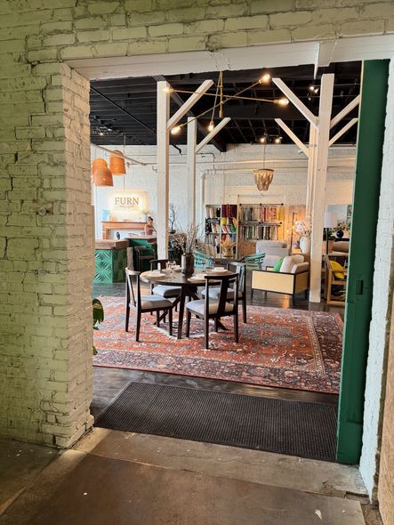 View through a doorway into a store. Dining table and furniture on patterned rug, white brick, and exposed beams.