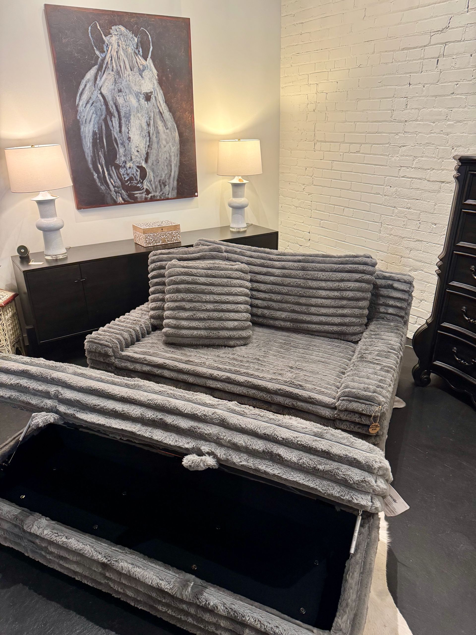 Gray textured sofa with two pillows, in a room with art, lamps, and a storage ottoman.