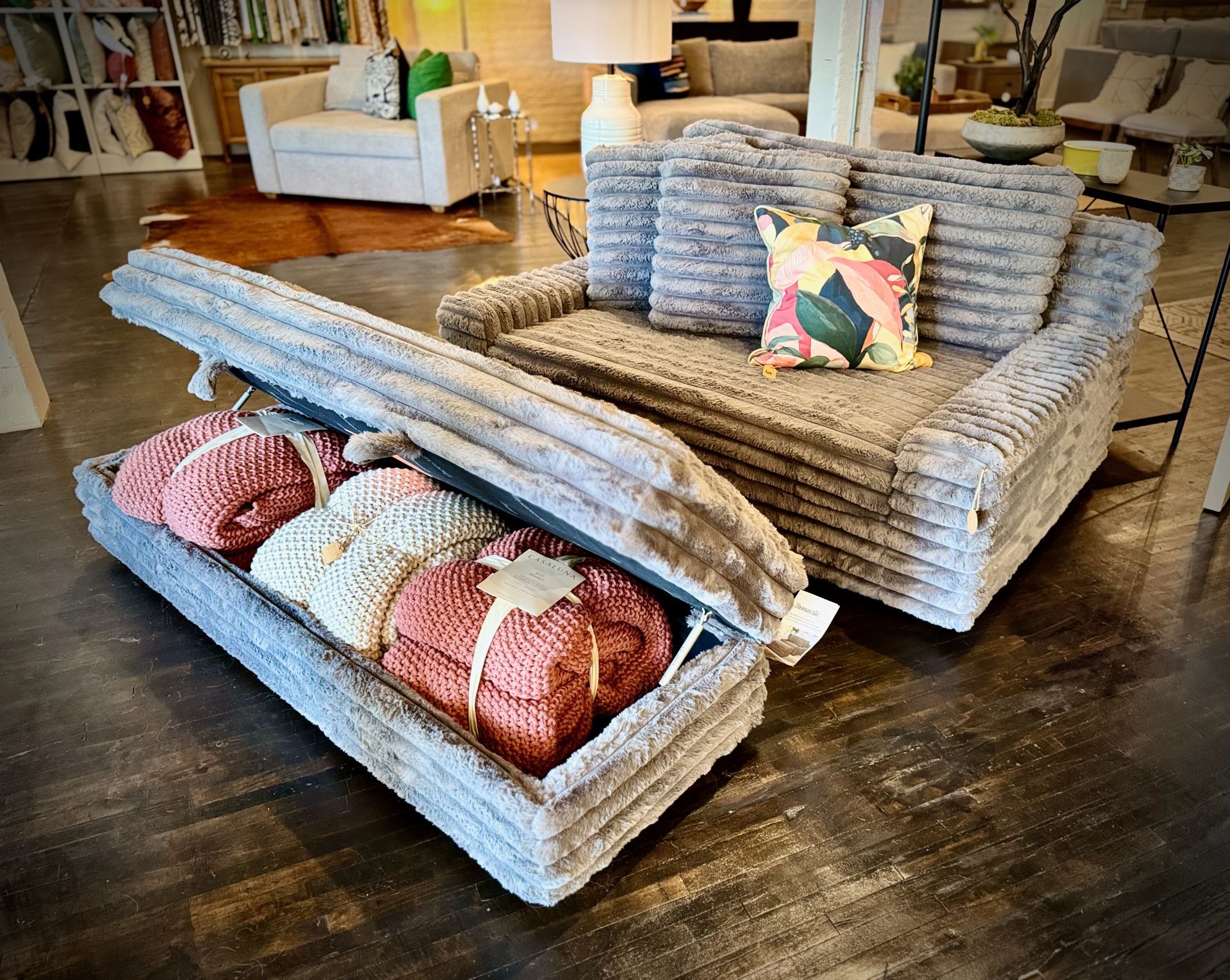 A gray faux fur storage ottoman and matching loveseat in a showroom, with blankets in the ottoman.