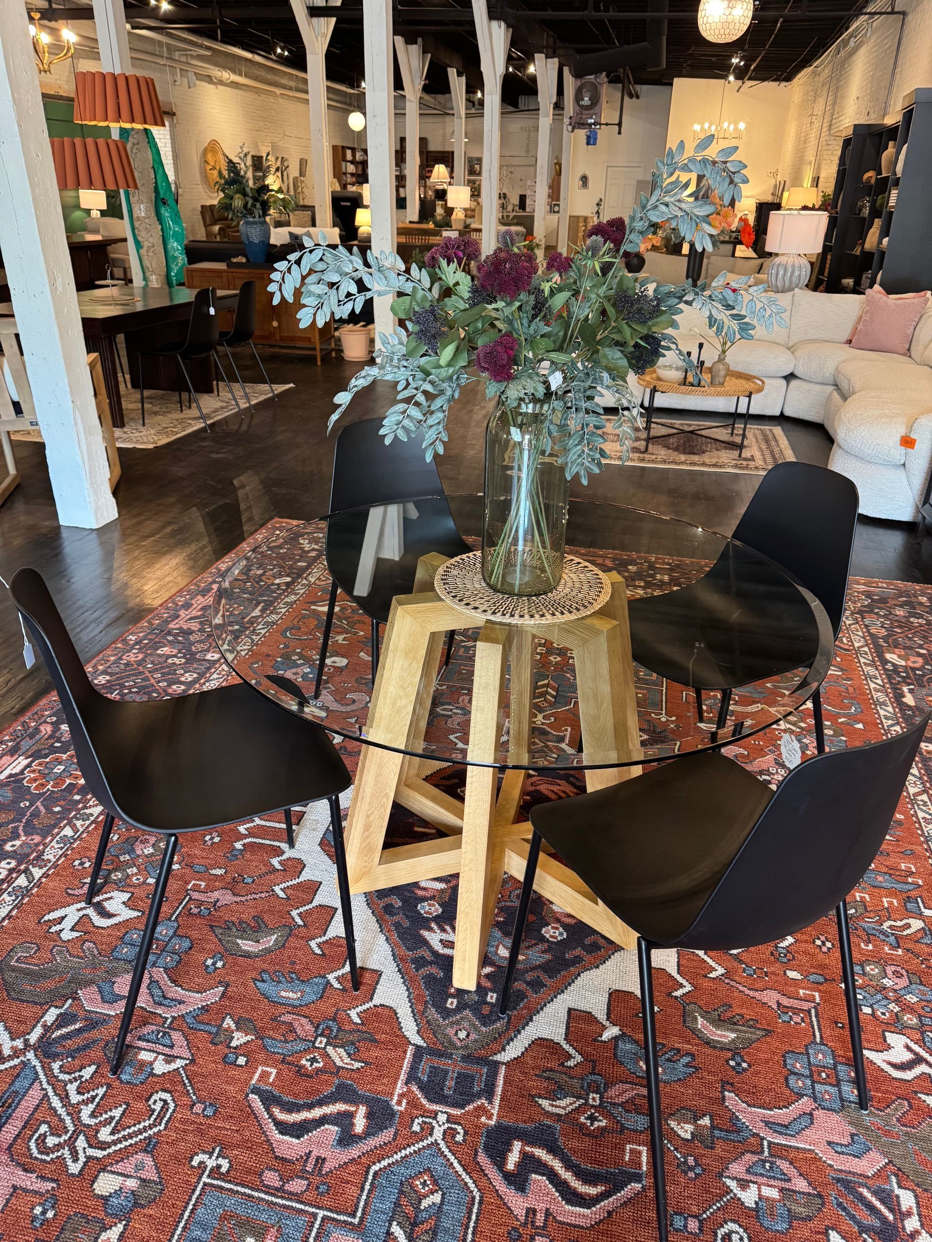 Dining area with black chairs around a wooden table, on a patterned rug, in a well-lit showroom.