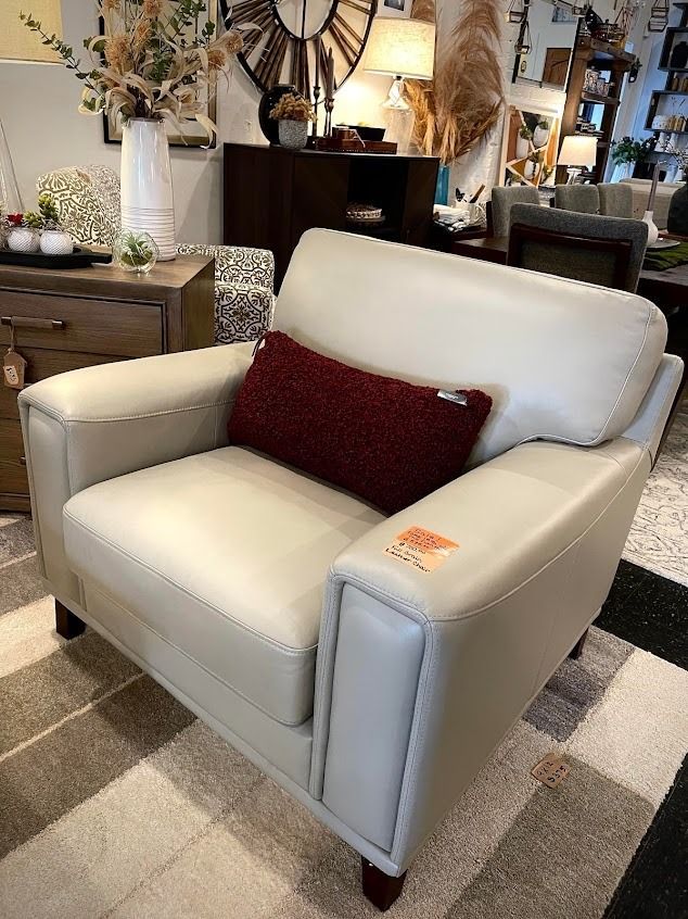 Cream-colored leather armchair with a red throw pillow in a furniture store, set on a checkered rug.