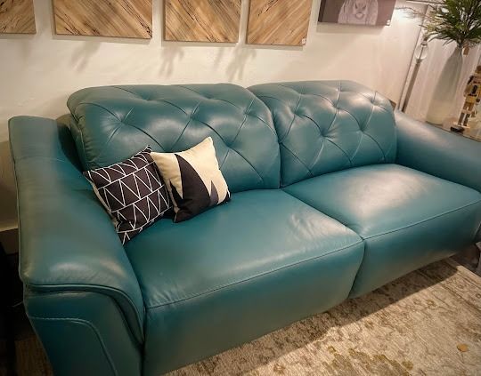 Teal leather sofa with two decorative pillows against a white wall with wooden art.