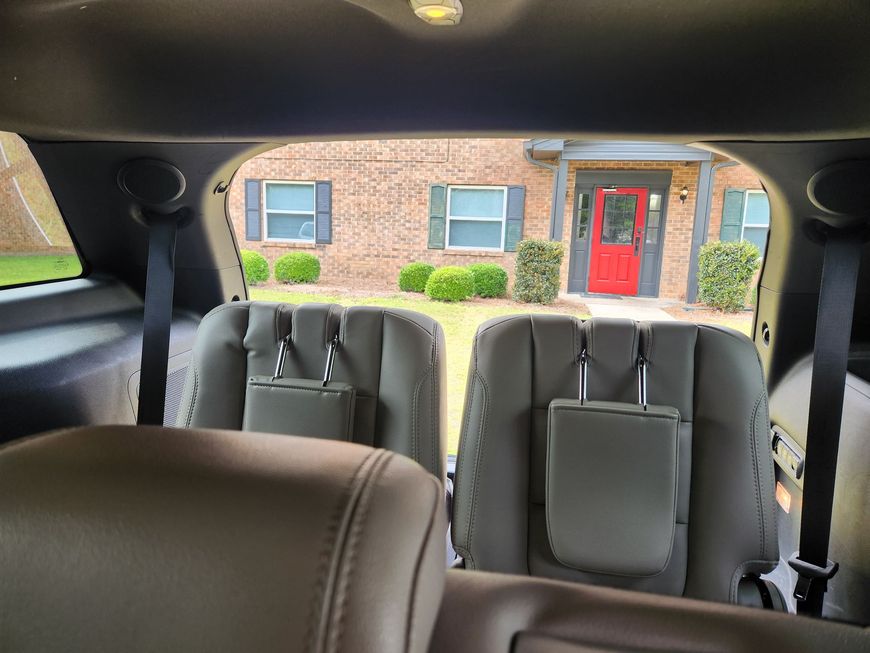 Interior of a car with gray seats, facing a brick house with a red front door through the rear window.