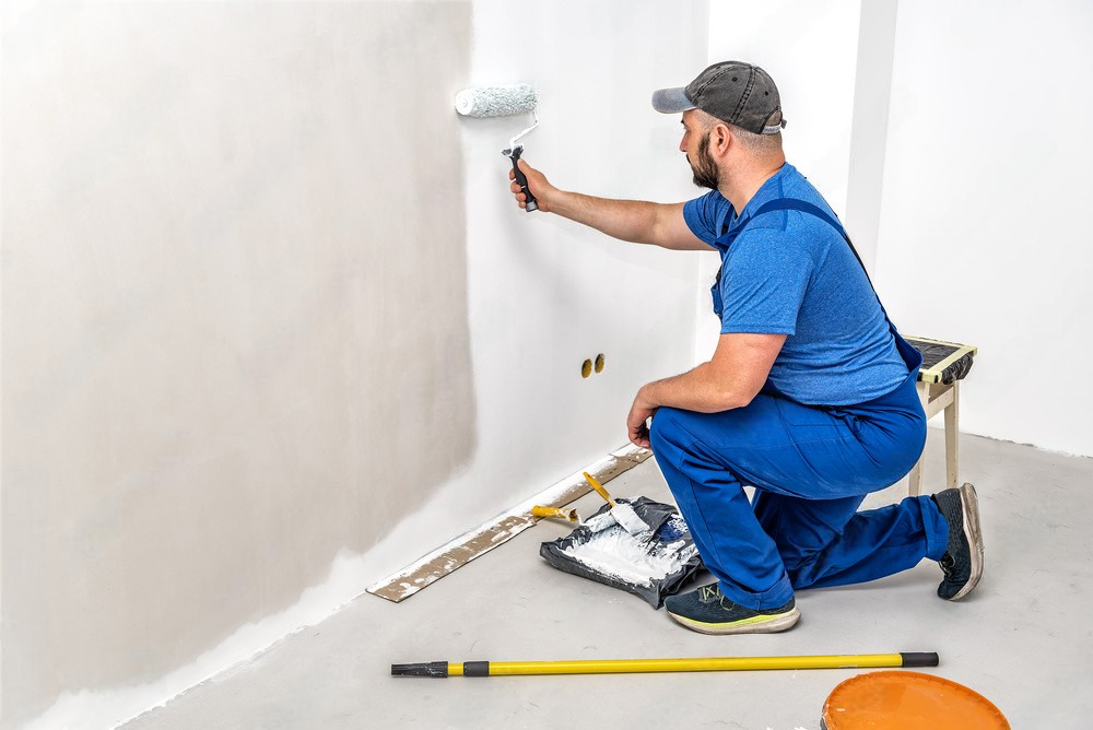 A man is kneeling down while painting a wall with a paint roller.
