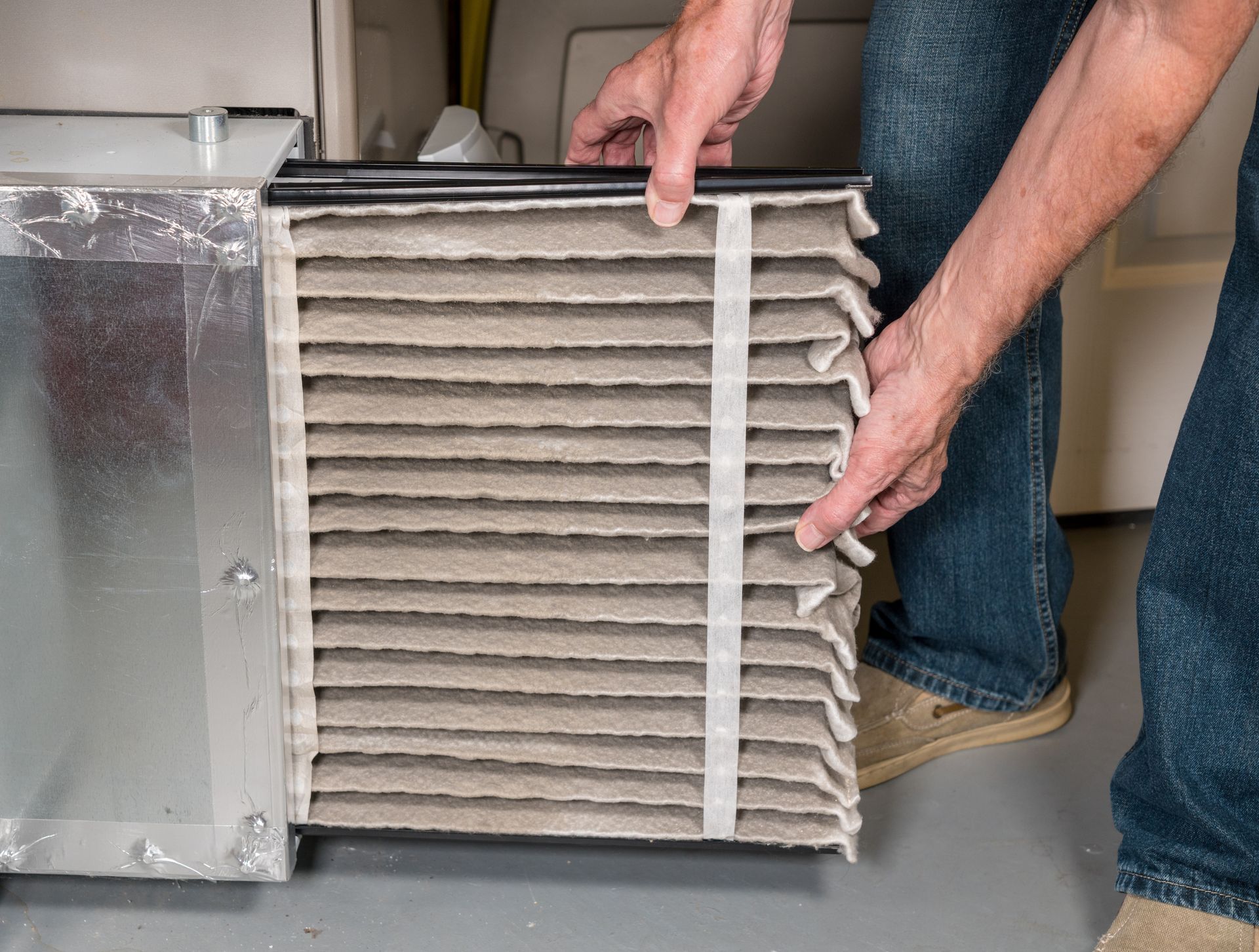 A person's hands holding a dirty air filter, preparing to replace it in an HVAC furnace.