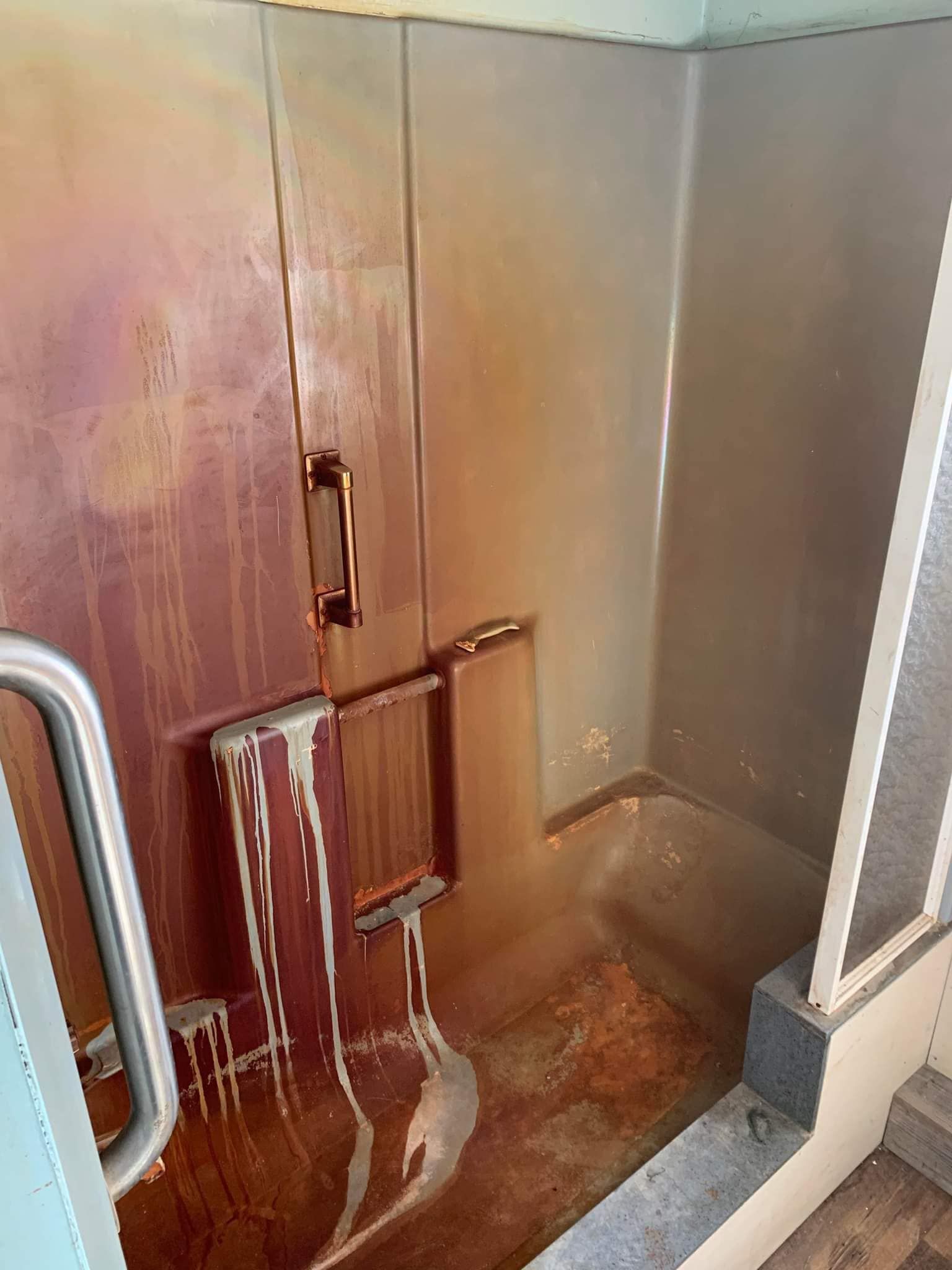 Rust-streaked stainless steel utility compartment with a door, pipes, and a wet floor