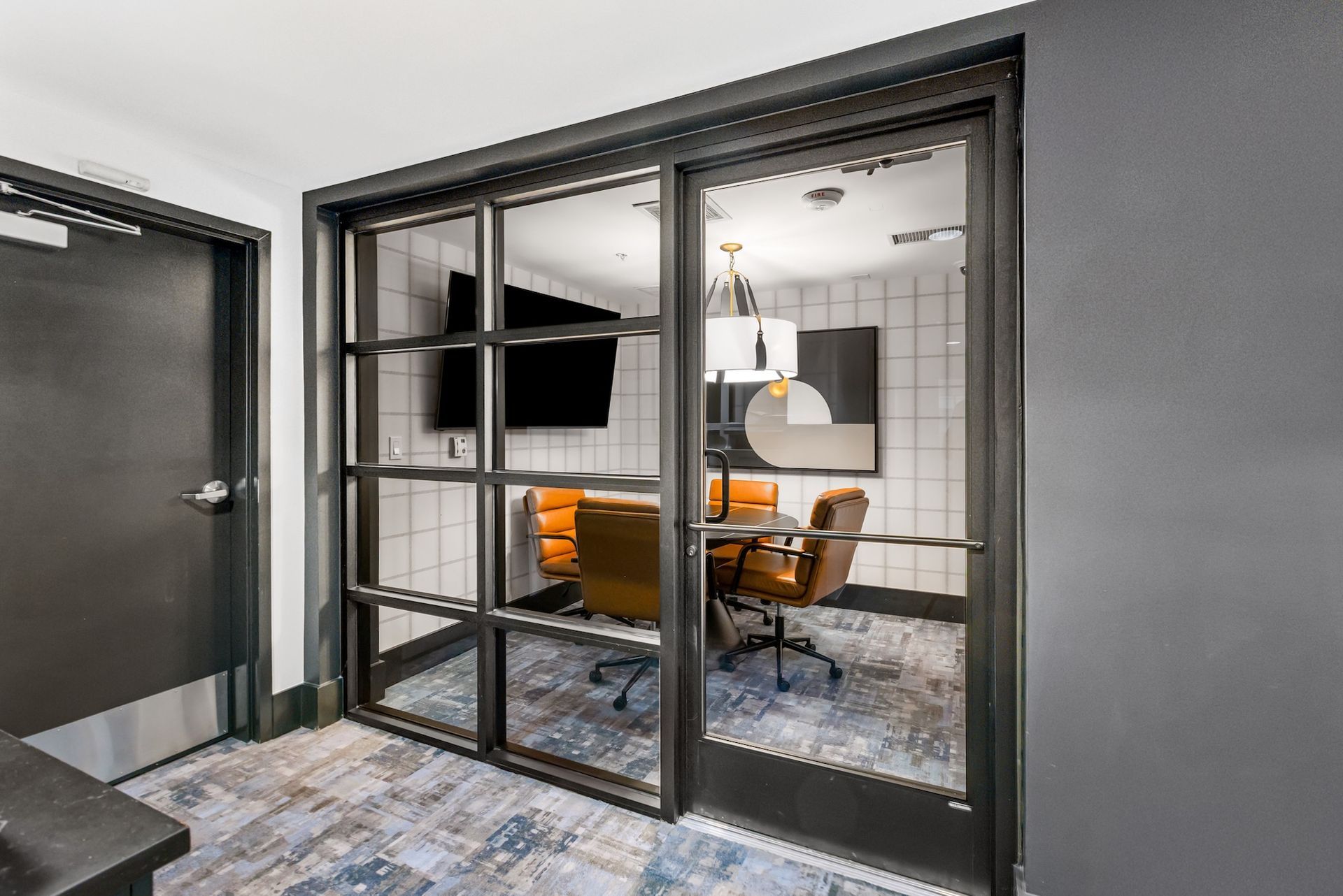 Glass-walled conference room with orange chairs, table, and a TV; next to a dark door.