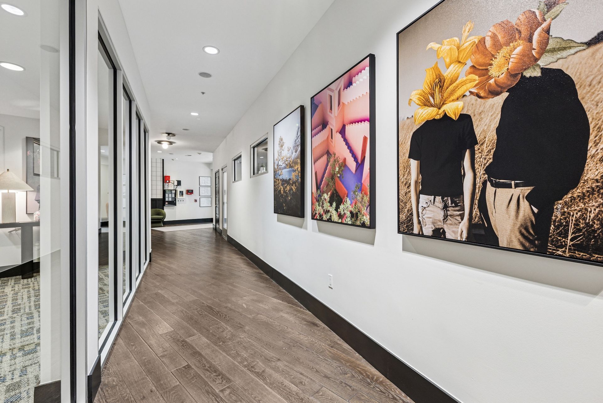 Long hallway with artwork on a white wall, glass-walled offices on the left, and wood flooring.