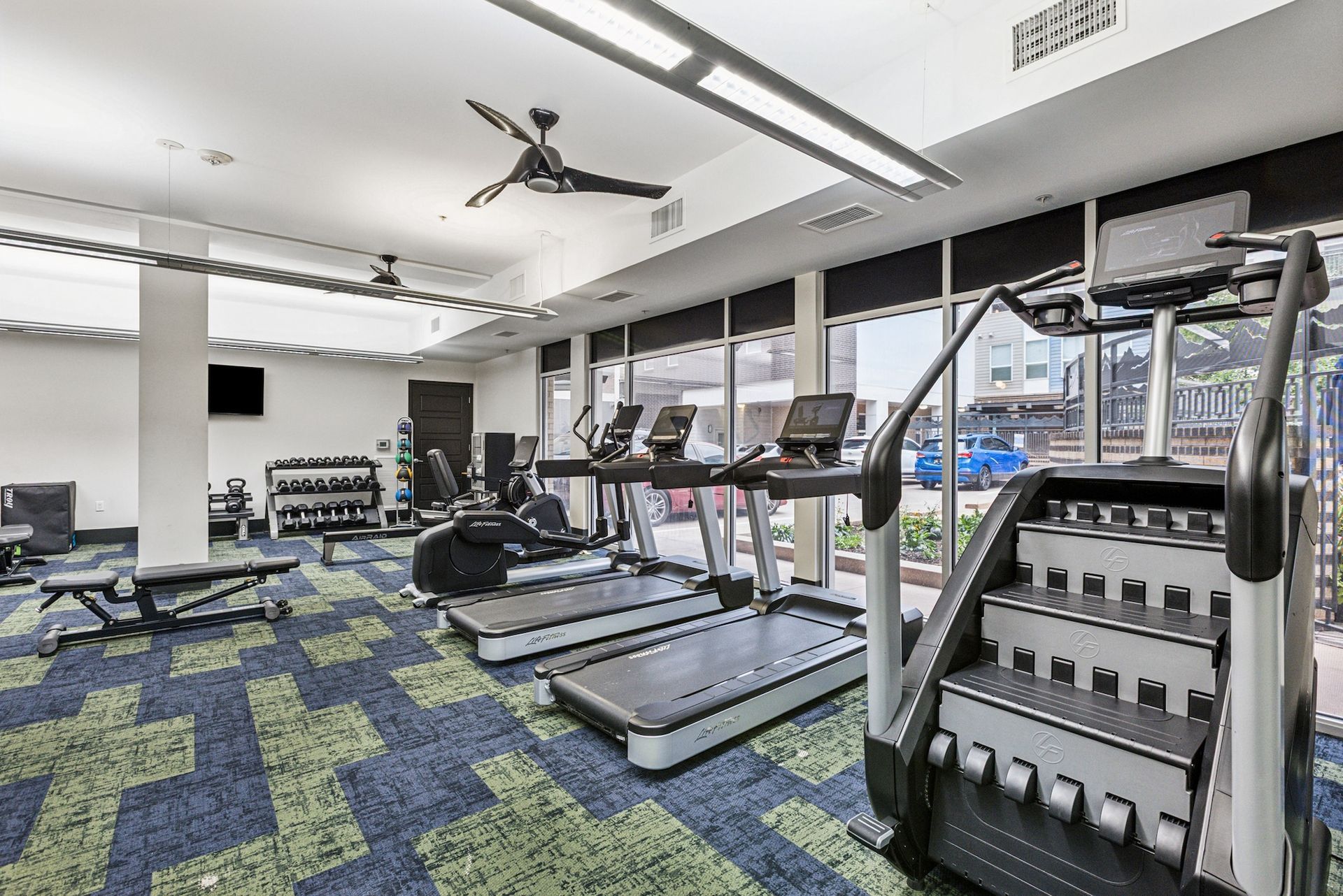 Gym interior with treadmills, stair stepper, weights, and blue/green carpet.