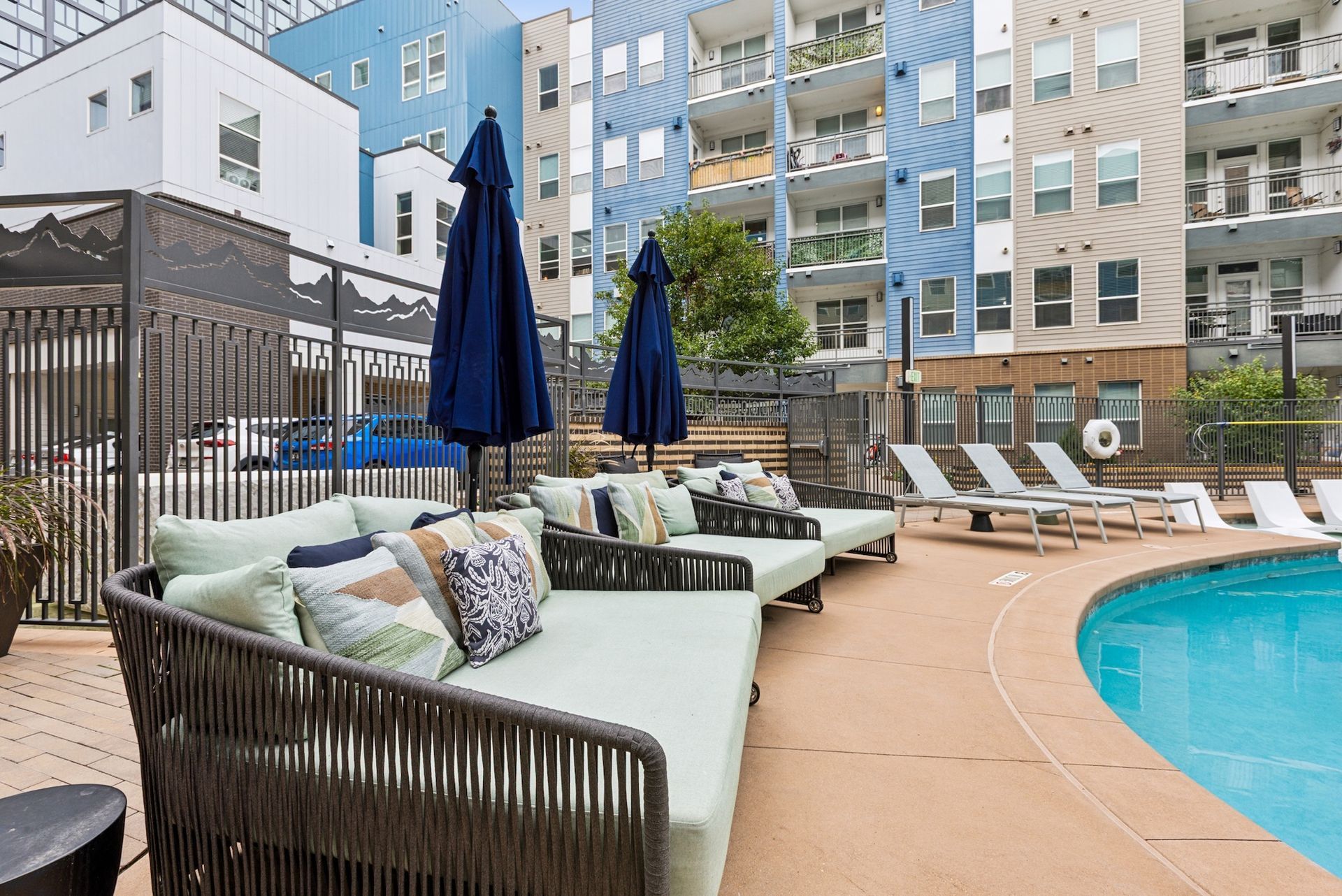 Poolside lounge area with blue umbrellas, chaise lounges, and a modern apartment building.