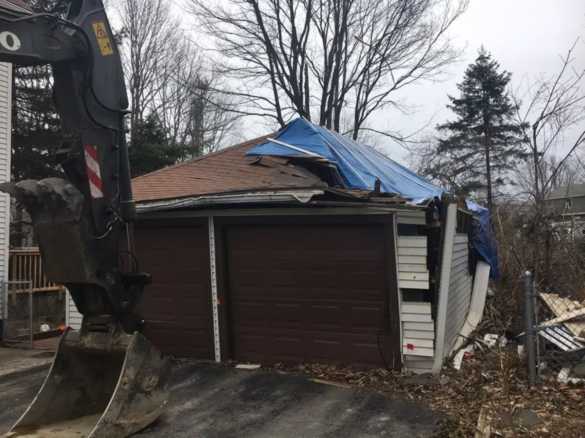 An excavator is demolishing a garage with a blue tarp on the roof.
