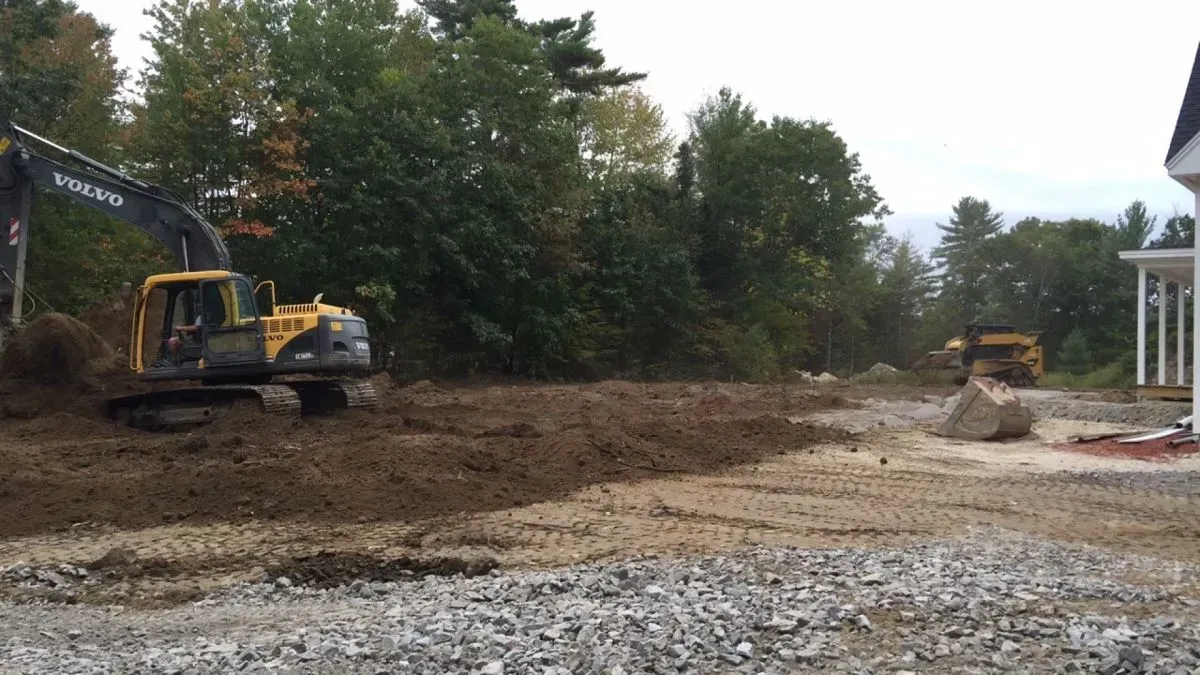 A volvo excavator is working on a dirt road in front of a house.