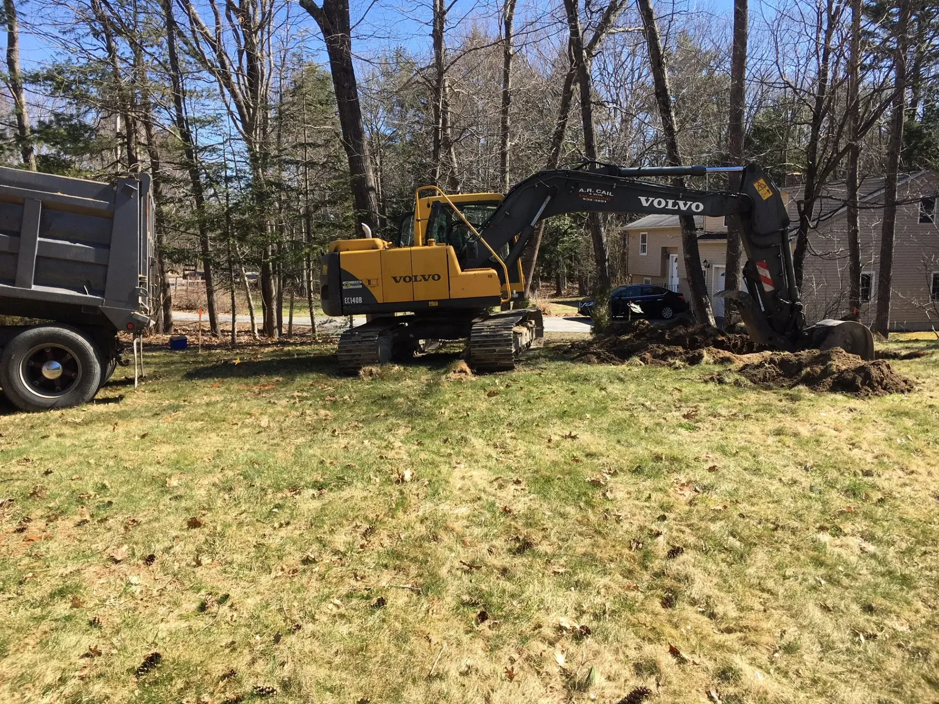 A yellow excavator is sitting in a grassy field next to a dump truck.
