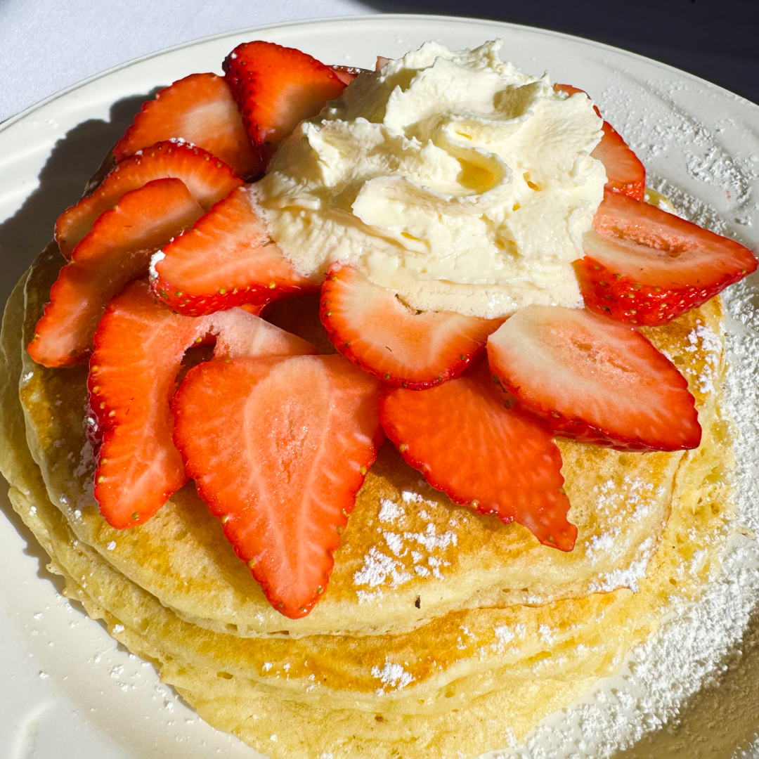 Fluffy stack of buttermilk pancakes topped with fresh sliced strawberries and a swirl of whipped cream, photographed in natural morning light.