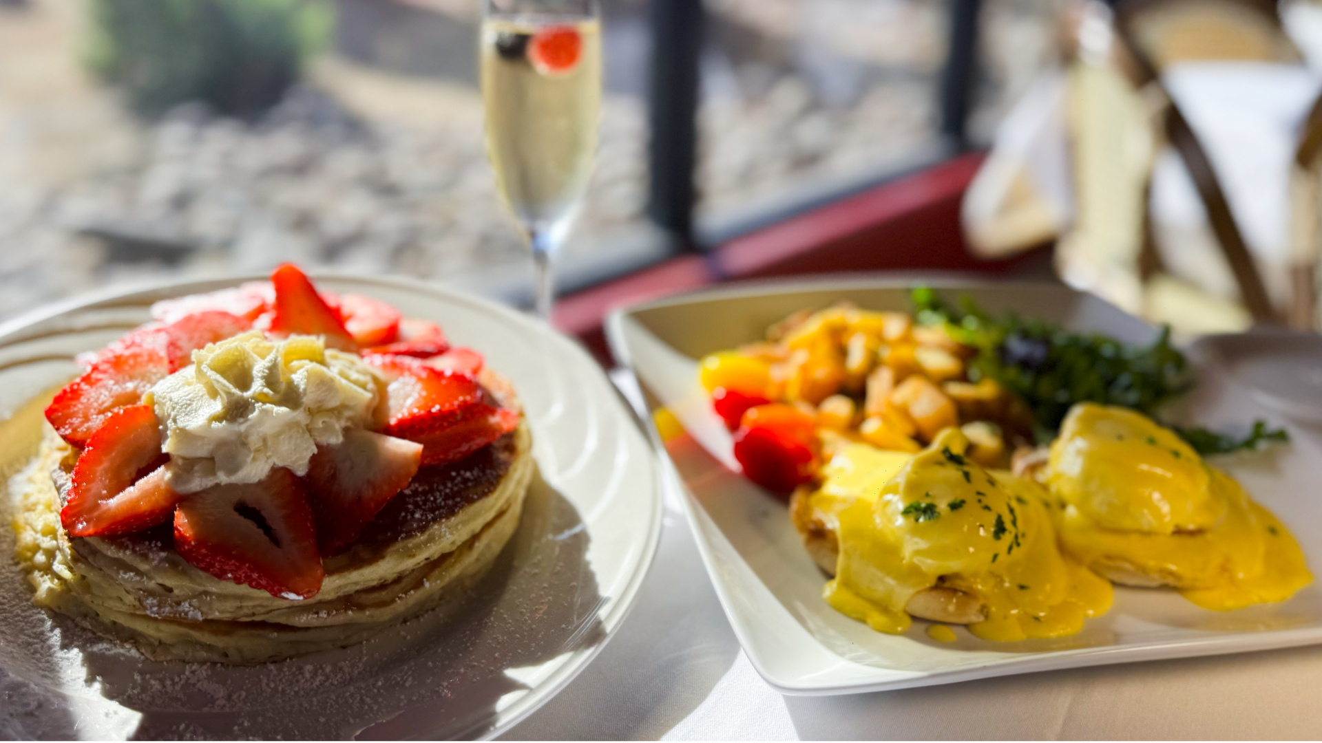 A beautifully plated French brunch at Chez Pierre Bistro featuring strawberry-topped pancakes, Eggs Benedict with golden hollandaise, crispy potatoes, fresh greens, and a glass of sparkling wine, photographed in natural morning light near the dining room window.