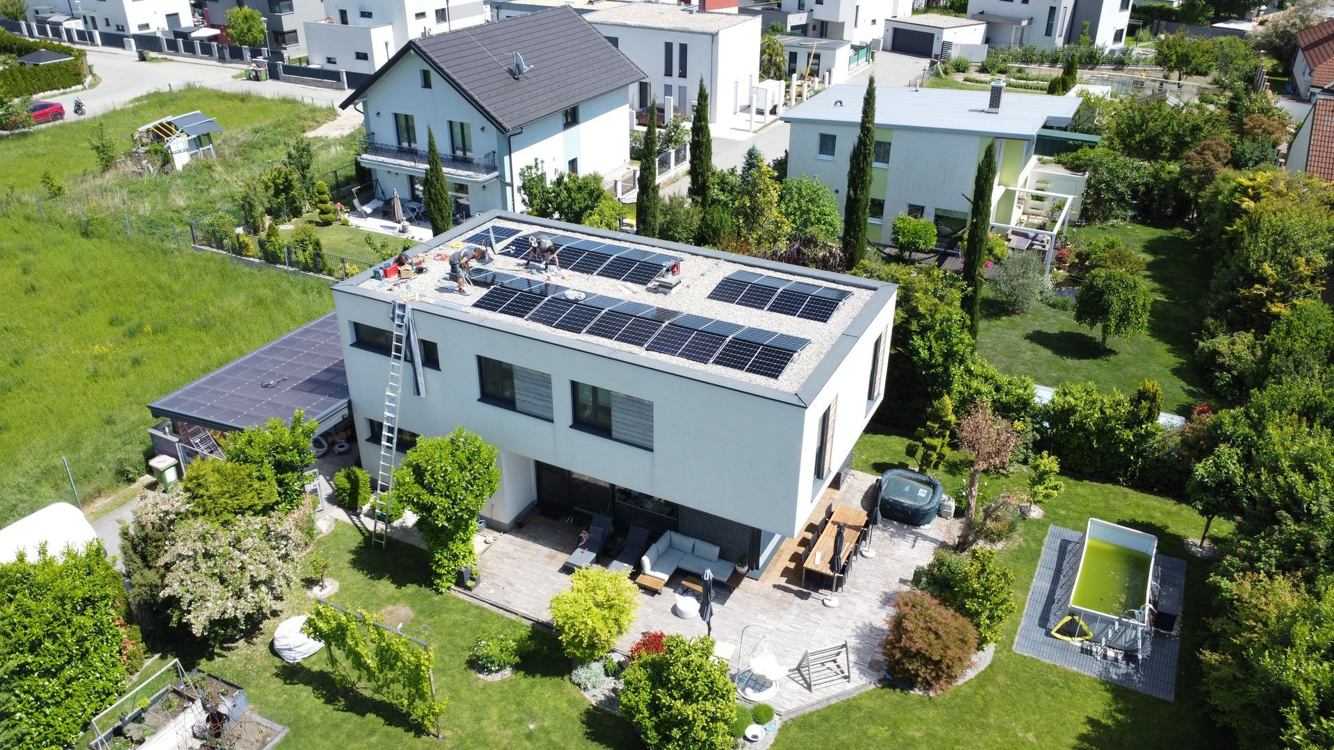 Luftaufnahme eines modernen weißen Hauses mit Solarpaneelen, umgeben von einem grünen Garten und Nachbarhäusern.