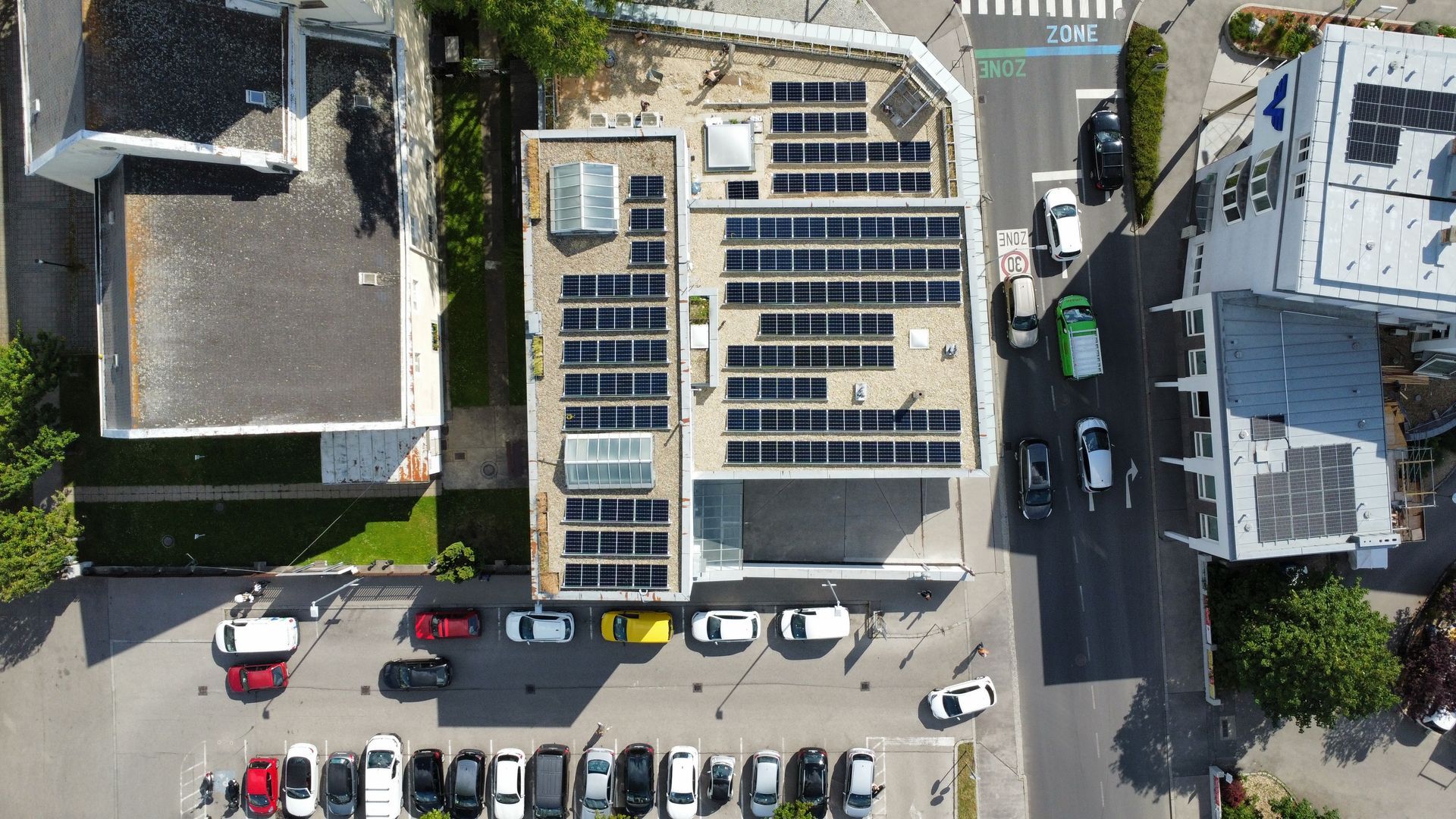 Luftaufnahme eines Gebäudedachs mit Solarpaneelen und geparkten Autos auf der darunterliegenden Straße.