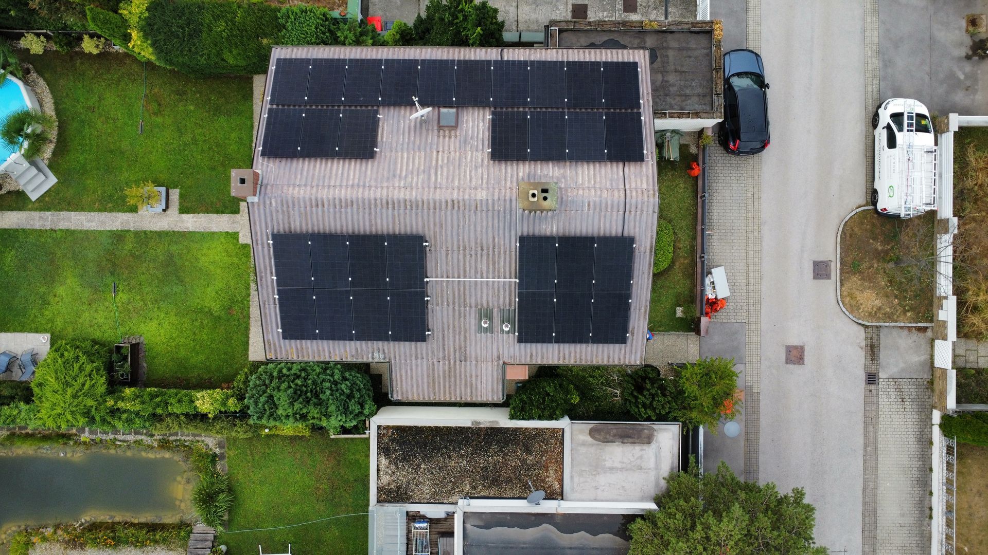 Luftaufnahme eines Hauses mit Solarpaneelen auf dem Dach neben einer Straße und geparkten Autos.
