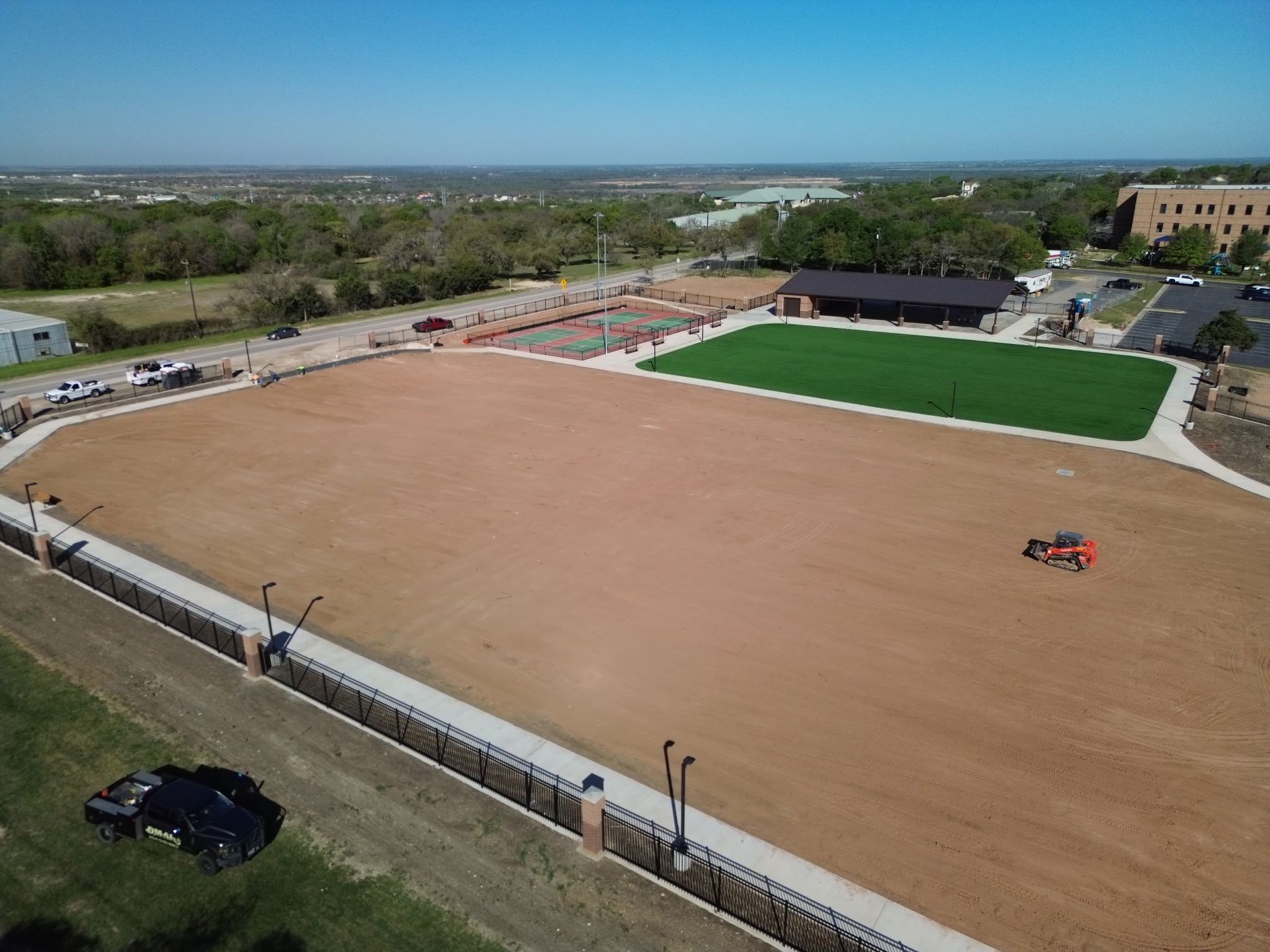 Aerial view of a large dirt sports field with a green turf area and small groups near the edge