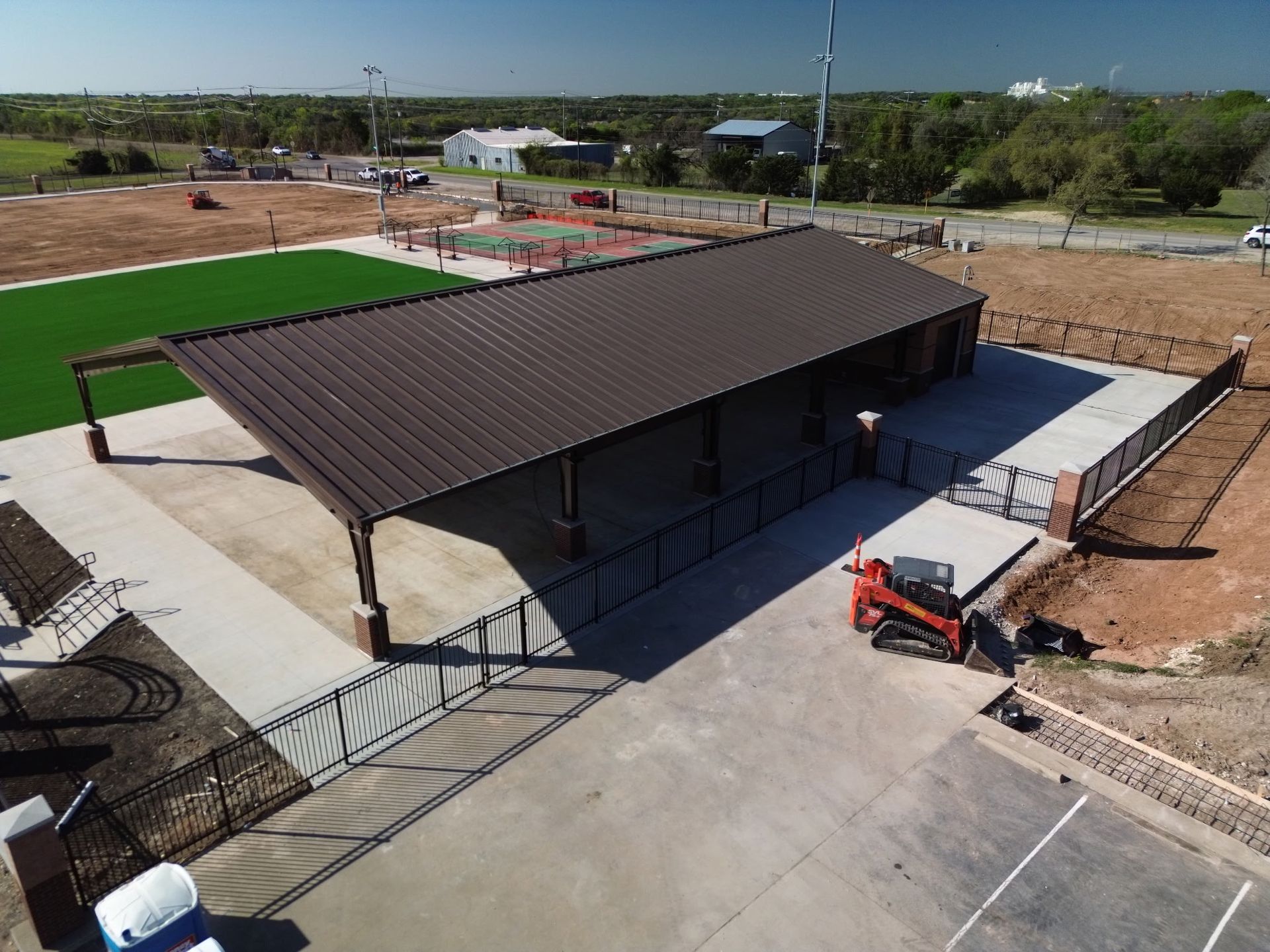 Aerial view of a pavilion with a dark roof beside a concrete lot and a small construction vehicle.