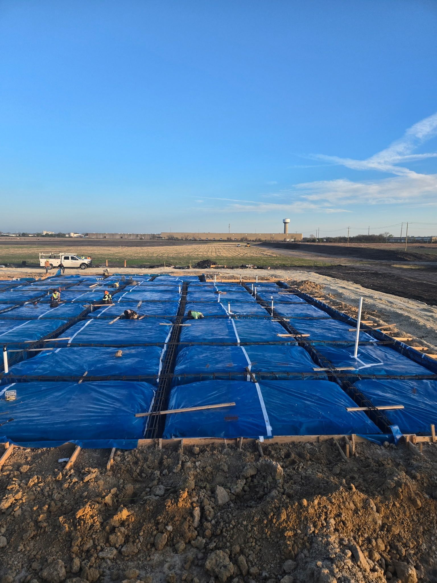 Blue tarps covering a rectangular field under a clear sky, with dirt foreground and distant structures.