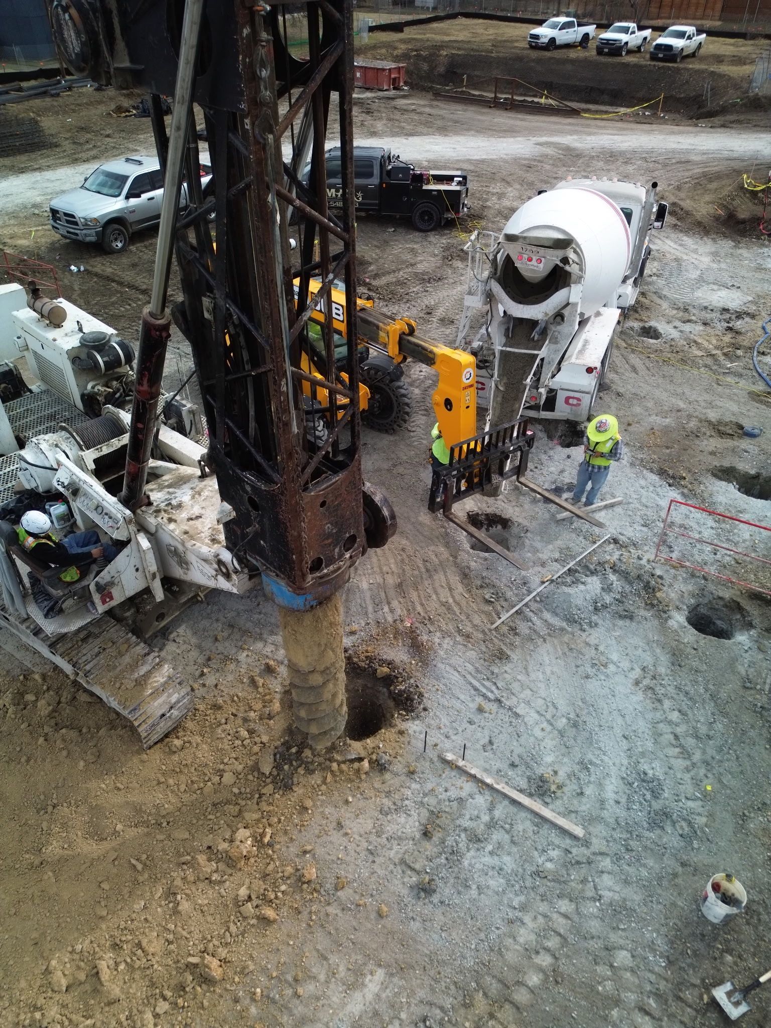 Construction site with drilling rig, concrete mixer truck, workers, and excavator on muddy ground