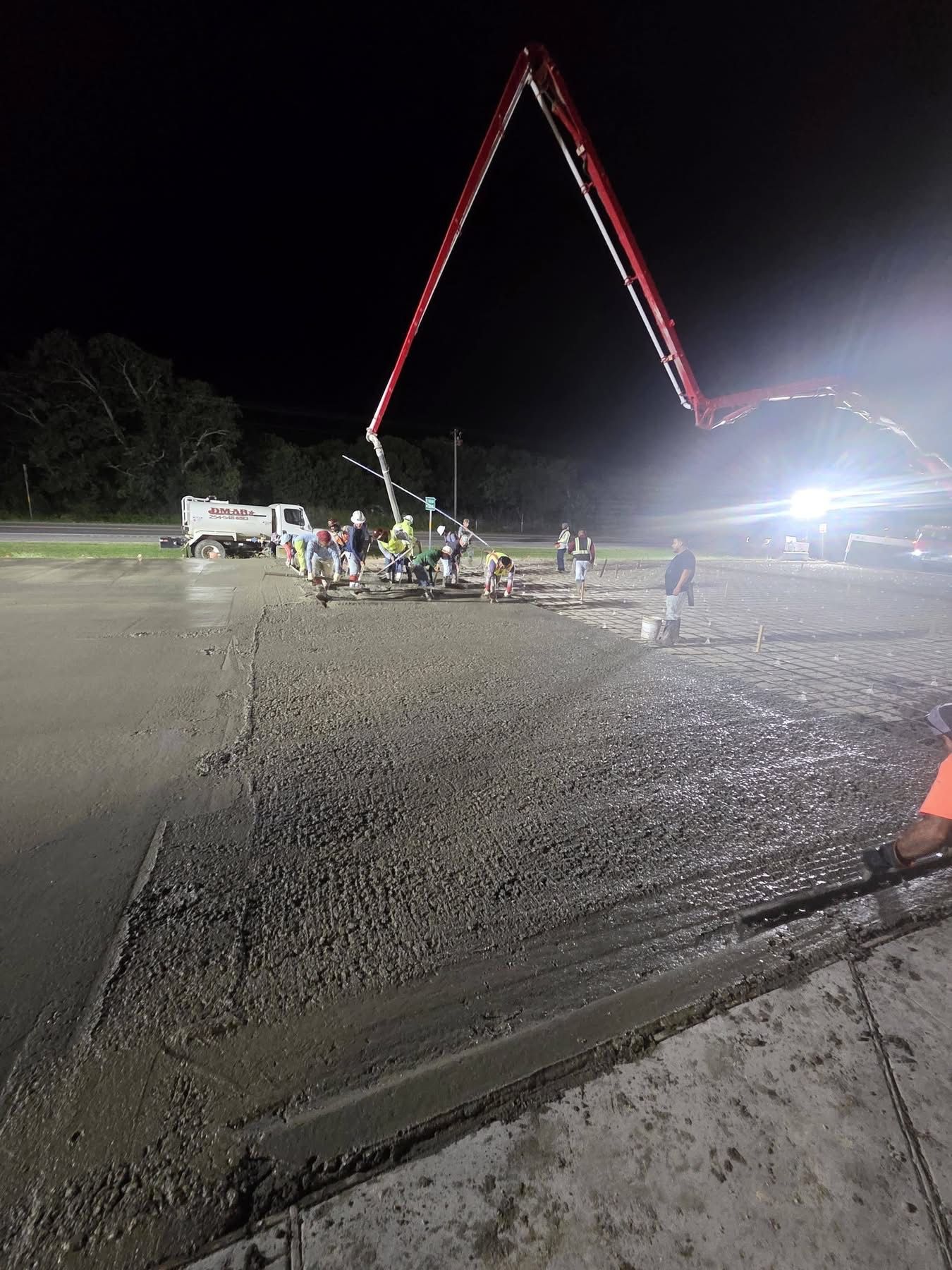 Night construction site with workers, a crane lit by red lights, and a bright spotlight over a gravel lot