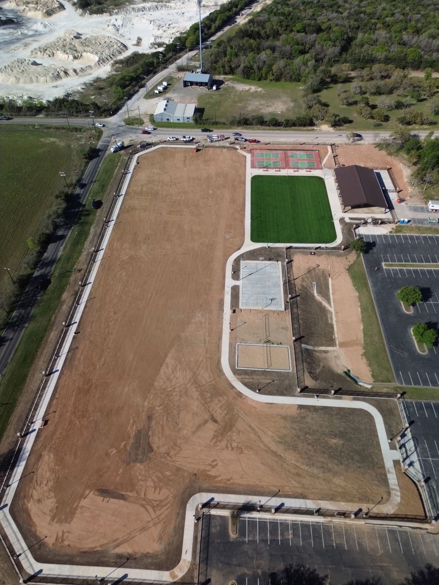 Aerial view of a sports field and dirt lot beside a paved parking area and road.