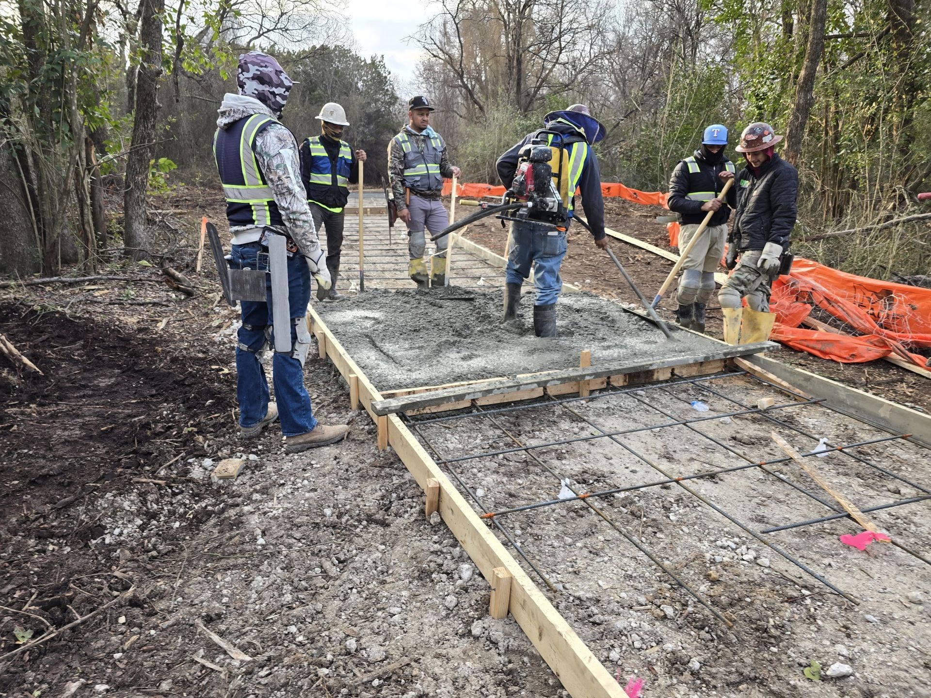 Construction workers pouring concrete for a walkway in a wooded area with orange safety fencing