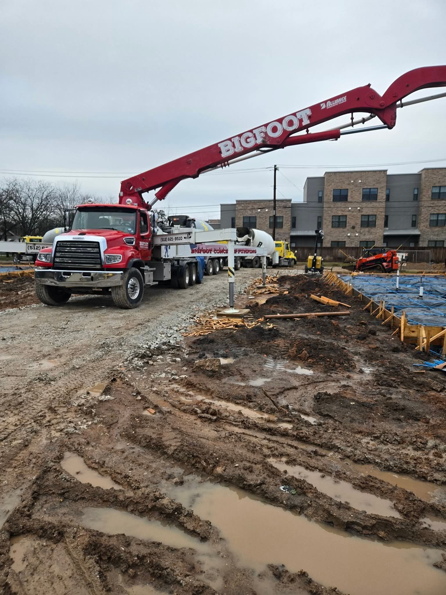 Red Bigfoot concrete pump truck at a construction site, muddy ground, boom extended.