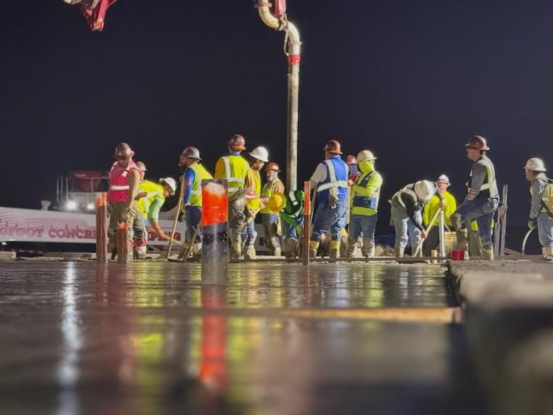 Construction workers pouring concrete at night. Workers wear safety vests and helmets, illuminated by lights.