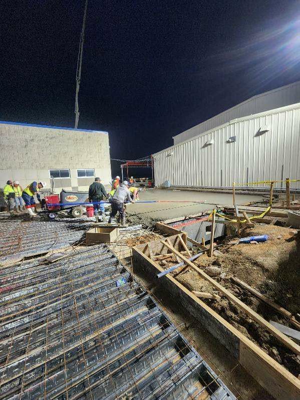 Construction workers pouring concrete at night. Rebar and wooden forms in foreground, buildings and a concrete pump in background.