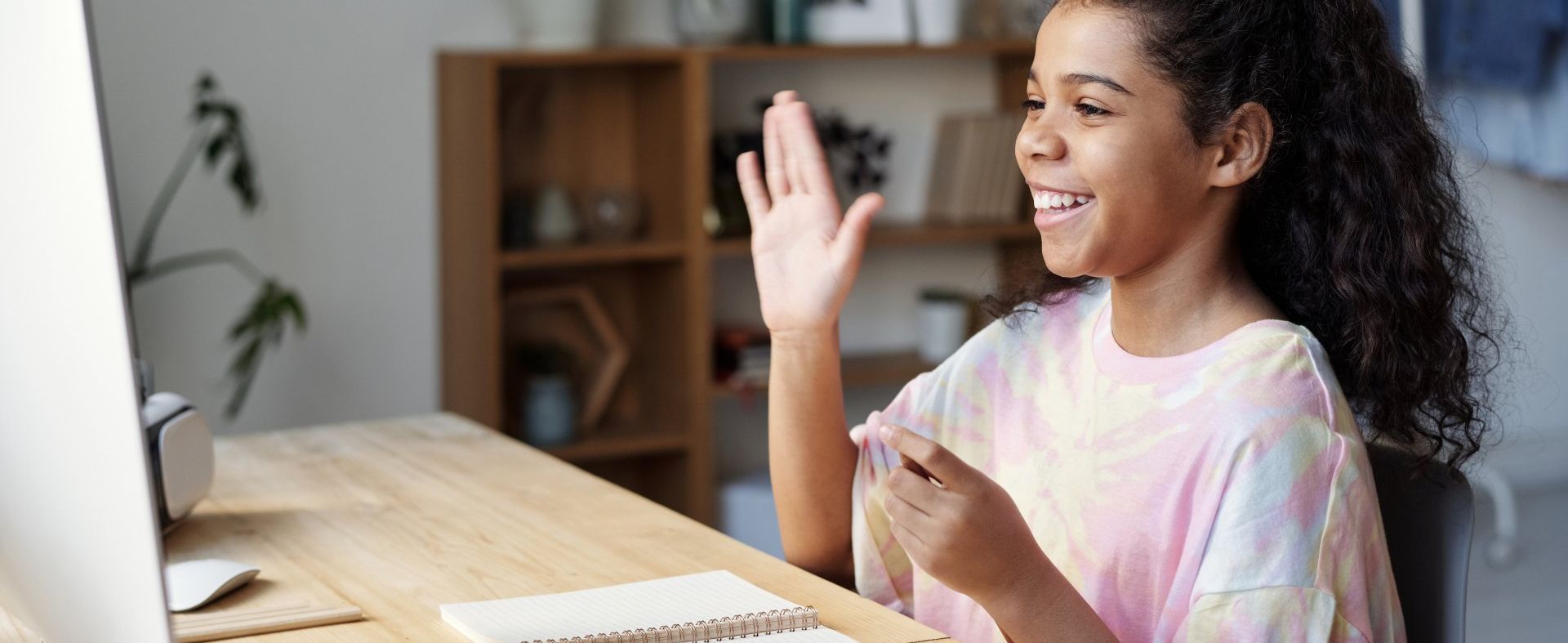 a young girl is sitting at a desk using a computer and waving at the camera .