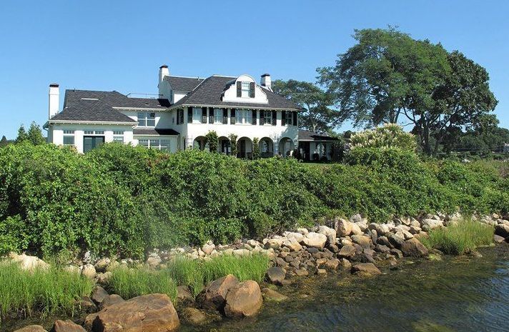 A large white house sits on a hill overlooking a body of water