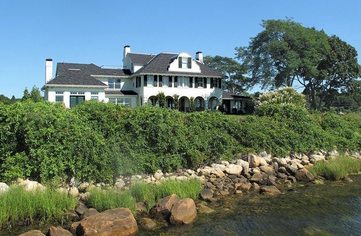 A large white house sits on a hill overlooking a body of water