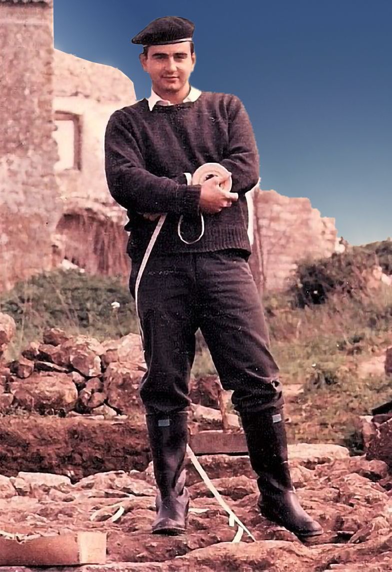 A man in a sweater and boots is standing in a field with his arms crossed.