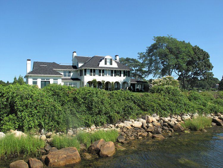 A large white house sits on a hill overlooking a body of water