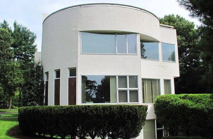 A white house with a curved roof is surrounded by bushes and trees